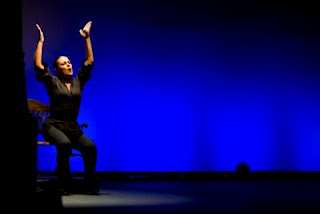 Mari Ángeles, brazos en alto sentada en el banco sobre un fondo azul