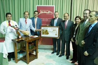 Junto a un jamón cortado, el consejero de Agricultura y Pesca de la Junta, el delegado territorial y el director en Huelva posan con la imagen del cupón dedicado al congreso mundial del jamón