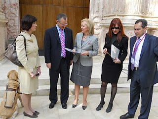 La presidenta del Parlamento sostiene en sus manos el orden del día en braille entregado por el delegado territorial de la ONCE en Andalucía junto al subdelegado y la presidenta y vicepresidenta del Consejo, a la entrada del salón de plenos