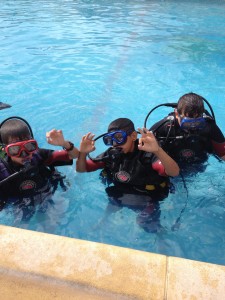En la imagen, tres niños vestidos de buzo saludan a la cámara desde el agua de la piscina