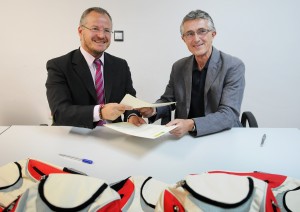 El delegado territorial de la ONCE y el presidente de la Asociación de la Prensa de Sevilla muestran a la cámara el contrato de colaboración mientras lo firman. Junto a ellos algunas d elas mochilas enviadas a Guinea-Bissau