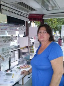 En la imagen, la vendedora María Aguilar mira a cámara desde dentro de su kiosco ONCE. En él aparecen de fondo cupones de la ONCE