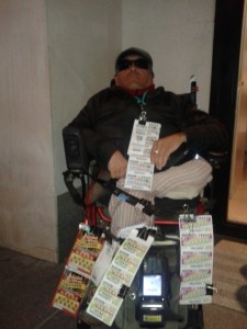 Carlos Vidal está en una silla se rueda, con una pierna amputada. Tiene los cupones sujetos a la silla y a la ropa