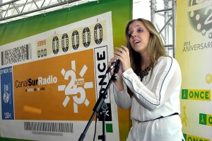 Se puede ver a Coraluna vestida de blanco, congiendo el micro con las manos. De fondo, el cupón naranja dedicado al 25 aniversario de RTVA
