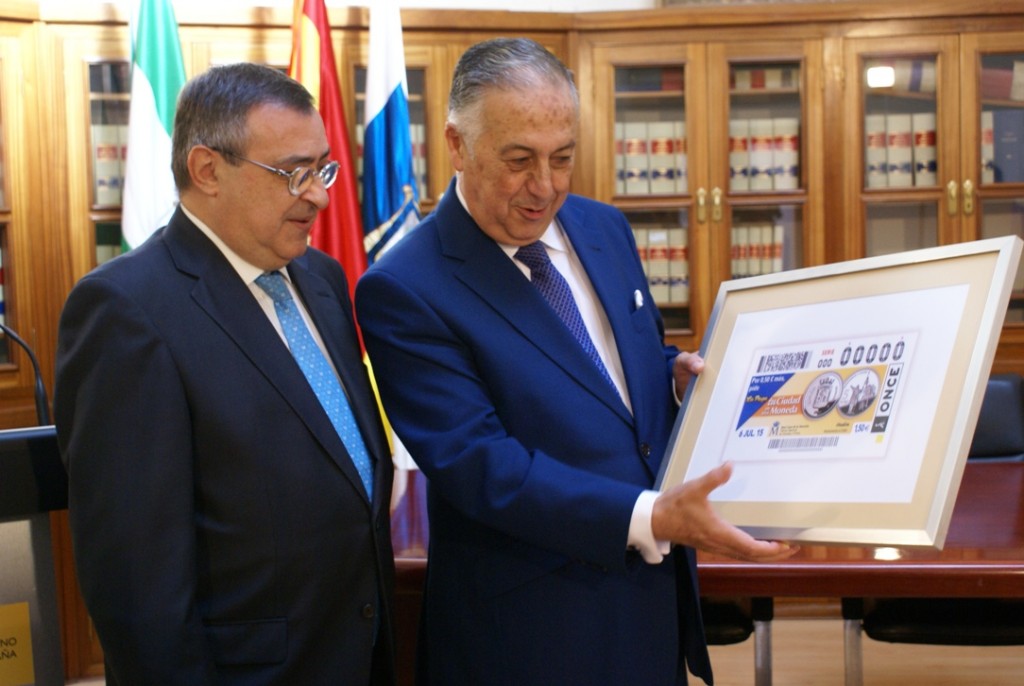 El subdelegado del Gobierno en Huelva y el director de la ONCE en la ciudad muestran el cupón de la moneda de Huelva, que pone como protagonista el monumento de Colón señalando América