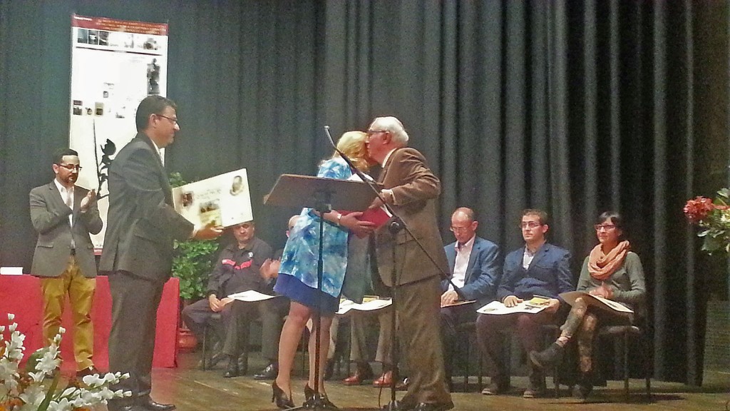 Carmen Aquilera, en el escenario, recibe el premio Quijote 2016 de manos del alcalde de Baena y cruzan un amistoso saludo mientras el resto de asistentes aplauden