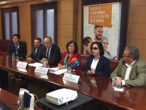 Rodeada por el resto de integrantes de la campaña solidaria y sentados a la mesa donde expusieron los preceptos de la campaña, Isabel Viruet toma la palabra mientras es observada por algunos de sus acompañantes a la mesa. Tras esta, en el fondo de la sala se halla el cartel publicitario elaborado para la ocasión. 